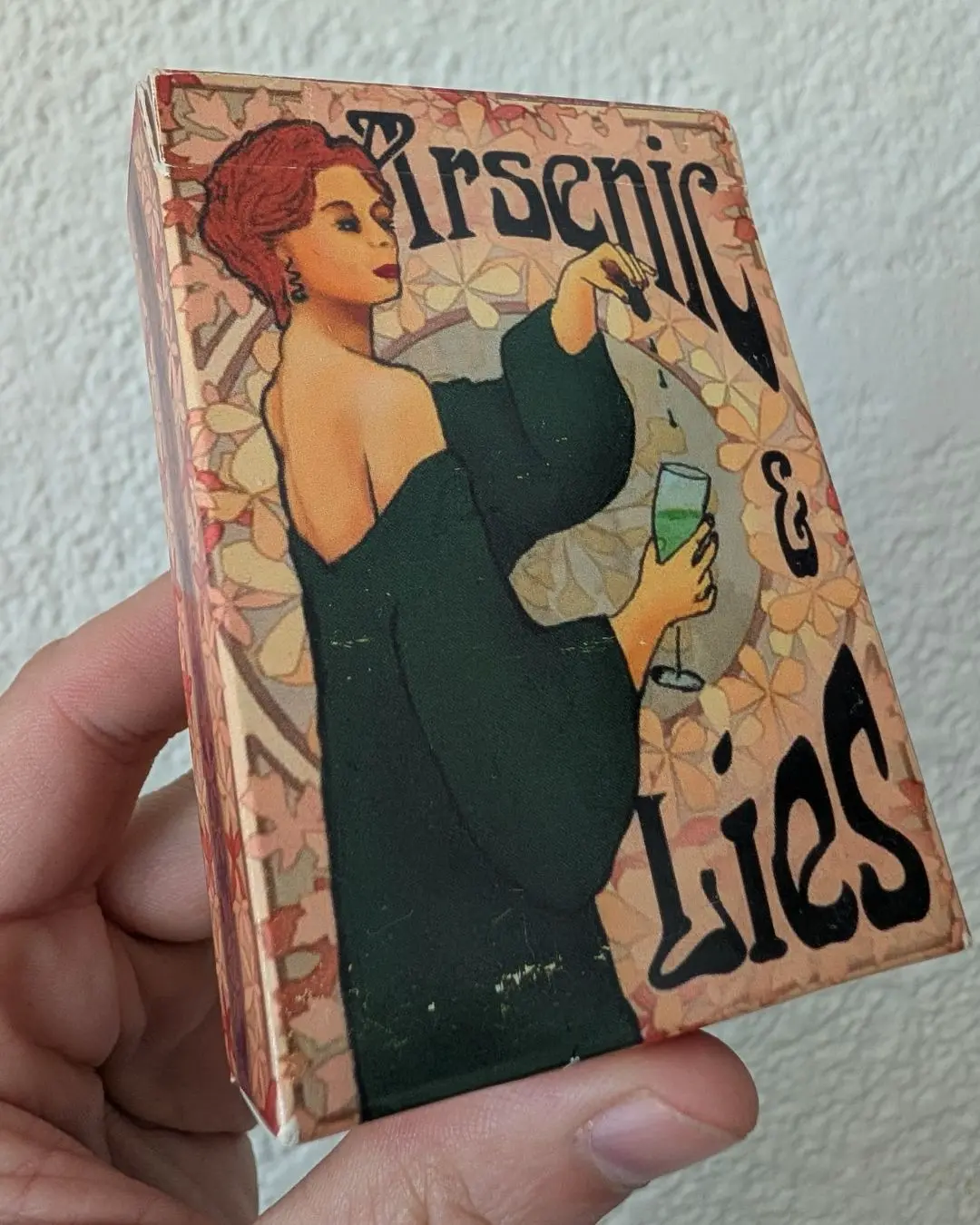 The Arsenic and Lies box held in one hand: an art-nouveau woman in a green gown tipping poison into a green cocktail.